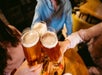 Three people clink glasses of beer over a wooden table with food, viewed from above in a casual indoor setting.