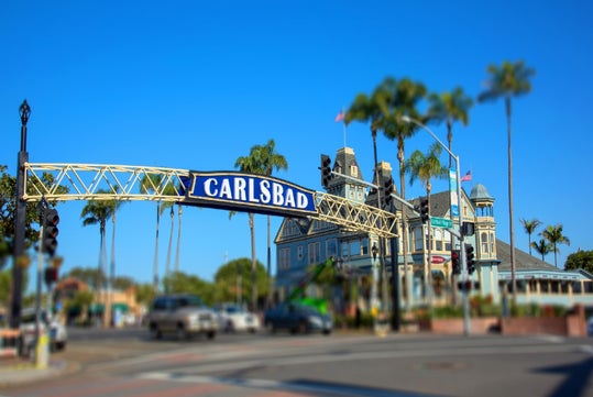 A large blue sign reading "Carlsbad" spans across a street, with traffic, palm trees, and a historic building in the background under a clear blue sky.