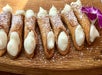A row of small cannoli filled with cream, dusted with powdered sugar, displayed on a wooden board with a purple flower garnish.