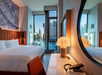 Contemporary hotel room with a large bed, a round mirror, a desk lamp, and a cityscape view through large windows.