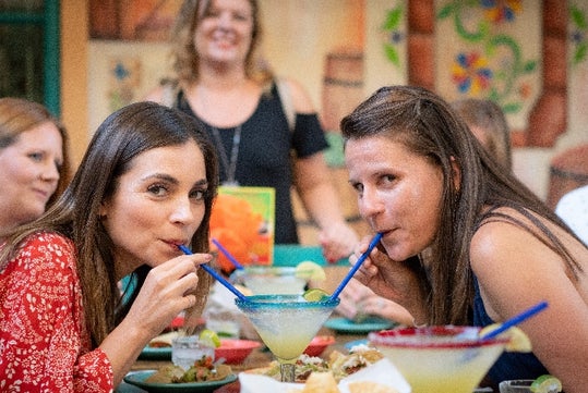 Two women sit at a table drinking from the same margarita glass with blue straws, surrounded by food, with other women in the background.