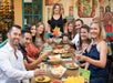 Nine adults sit around an outdoor restaurant table, smiling and raising drinks, with various Mexican dishes and chips on the table. A woman stands behind holding a menu.