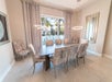 A modern dining room with a long rectangular table, eight upholstered chairs, two round pendant lights, large window with curtains, and a wall mirror.