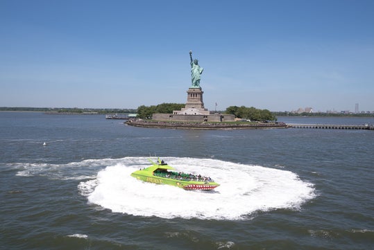 A green speedboat labeled "BEAST" makes a circle in the water near the Statue of Liberty, with the New York City skyline in the background.