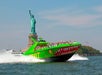 A green speedboat with a shark design speeds past the Statue of Liberty on a clear day.