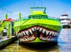 A green boat with a painted shark mouth design on the front, docked at a pier with another boat in the background.