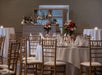 A banquet room with white cloth covered tables set for dinner with floral center pieces and wooden chairs with white cushions.