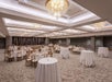 A large ornate white conference room with several white cloth covered standing tables, large white cloth covered tables set for dinner, and chandeliers overhead.