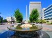 A round fountain with cascading water in the foreground, flanked by two trees and with a tall obelisk in the background, surrounded by buildings under a clear blue sky.