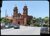 A brick church with two domed bell towers stands beside a road with cars and bicycles, under a clear blue sky.