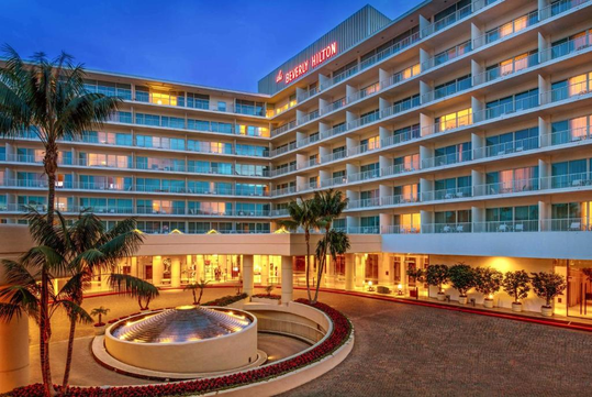 A multi-story Hilton hotel building at dusk, with illuminated windows, palm trees, and a circular driveway featuring a fountain in front.