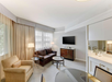 Bright hotel room with a sofa, armchair, coffee table, and wall-mounted TV. Large windows with sheer curtains let in natural light, and there's a kitchenette with a counter and chairs.