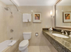 Hotel bathroom with a bathtub, toilet, and granite countertop. Towel hangs on wall rack, framed artwork above toilet. Sconce lighting and large mirror above sink.