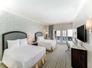 Hotel room with two double beds, striped carpet, a wall-mounted TV, desk, and a balcony with a pool view. Bright lighting and patterned wallpaper create a spacious atmosphere.