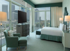 A hotel room with a large bed, armchair, TV, and windows offering a cityscape view. The decor is in soft blue and green tones.