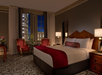 Elegant hotel room with large bed, red accents, two armchairs, and a city view through a window. Nighttime scene with warm lighting and classic decor.