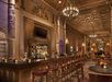 Elegant bar with wooden decor, ornate ceiling, chandeliers, and a television. Rows of brown chairs line the bar with various bottles displayed on shelves. Tables are set with small lamps and flowers.