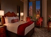 Hotel room with a large bed, red accents, two red armchairs, a bedside lamp, and a window overlooking a cityscape at night.
