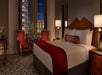 Hotel room with a large bed, red accents, and two chairs by the window. Night view of a lit building outside.