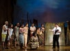 A group of people in colorful outfits stands on stage, facing a person in a white shirt holding a book, against a backdrop resembling a village setting.