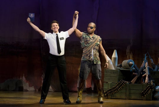 Two men on stage; one in a white shirt holds up a book and someone's arm, while the other is in military attire, surrounded by weapons and crates.