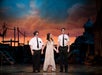 Three performers holding hands on stage, set against a dramatic sunset backdrop with rustic village scenery.
