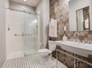 Bathroom with a glass-enclosed shower, white sink, and toilet. The wall has floral wallpaper, and the floor has a black and white tile pattern. Towels hang near the shower.