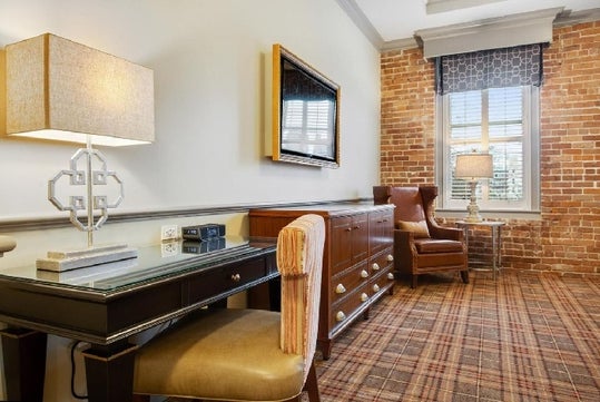 A stylish hotel room with a desk and lamp, a wall-mounted TV, a large armchair by a brick wall, and a window with patterned drapes.