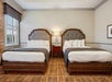 Hotel room with two double beds, a central lamp on a wooden nightstand, plaid carpet, and a window with patterned curtains on the left.