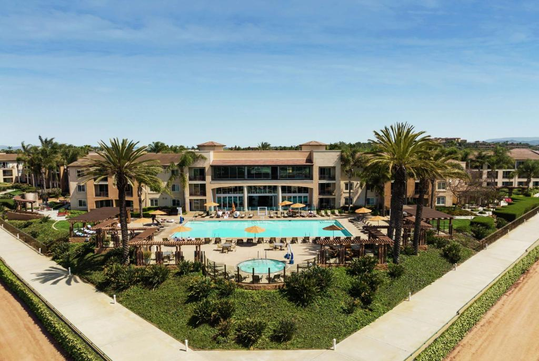 Aerial view of a hotel resort with a large central pool surrounded by palm trees, lounge chairs, and umbrellas. Pathways lead to the resort buildings under a clear sky.