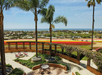 View from a terrace with palm trees, overlooking colorful flower fields and a distant ocean under a blue sky.