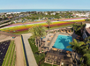 Aerial view of a pool area surrounded by palm trees and lounge chairs. In the background, colorful flower fields and a coastal landscape are visible.