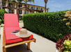 A red lounge chair with a hat and an open book is on a patio next to a green hedge. A building with balconies is in the background under a clear blue sky.