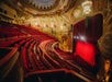 Ornate theater interior with red velvet seats and a large stage curtain, surrounded by elaborate architectural details and a grand ceiling.