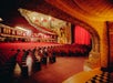 Empty ornate theater with red seats and a red curtain on stage. Archways and intricate designs decorate the ceiling and walls.