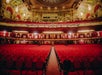 Empty theater auditorium with ornate ceiling, red seats, and balconies. The stage is not visible.