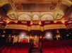 Ornate theater interior with red seating, gilded balconies, and decorative ceiling. Warm lighting enhances architectural details, creating an elegant atmosphere.