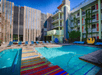Outdoor swimming pool area with colorful striped rugs, lounge chairs, and surrounding multi-story buildings under a clear blue sky.