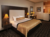 Hotel room with a king-sized bed, two bedside lamps, a desk with a chair, and patterned carpet. The room has warm lighting and a small sink area in the corner.