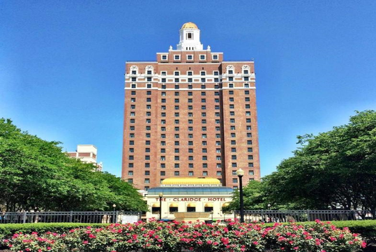 The image shows a tall, red-brick hotel building under a clear blue sky, with a dome at the top, surrounded by greenery and a foreground of blooming flowers.