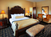 A luxurious hotel room with a large bed, patterned bedding, matching nightstands with lamps, an armchair set, a decorative bench, and a large mirror.