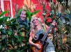 A man peeks through corn stalks on the left while a woman stands on the right, smiling and playing a guitar. They are in a colorful, farm-themed setting with red structures and greenery in the background.