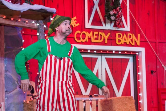 A performer in red and white striped overalls and a green shirt and hat stands on stage in front of a barn facade with the signage "The Comedy Barn." Lights and decorations are visible in the background.