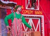 A performer in red and white striped overalls and a green shirt and hat stands on stage in front of a barn facade with the signage "The Comedy Barn." Lights and decorations are visible in the background.