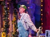 A person wearing a striped shirt, overalls, and a green cap balances a shoe on a striped stick held in their mouth onstage with string lights and sunflower decorations in the background.