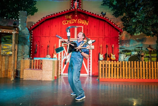 A performer in overalls juggles various items, including a toilet plunger and a shovel, on stage in front of a barn labeled "The Comedy Barn." Musical instruments are seen in the background.