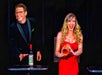 A man in a suit stands by a table with a bottle, while a woman in a red dress holds a cylinder, both appearing excited on a stage.