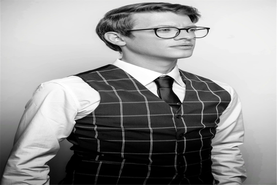 A person wearing glasses, a white shirt, and a checkered vest with a tie, gazing sideways. The image is in black and white.