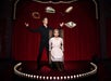 A magician performs on stage with a woman seated in a chair. Books and a tambourine hover above them. Red curtains and stage lights create a theatrical backdrop.