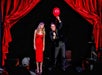 A woman in a red dress stands on stage next to a man holding a red balloon. They are under red curtains, performing to an audience.